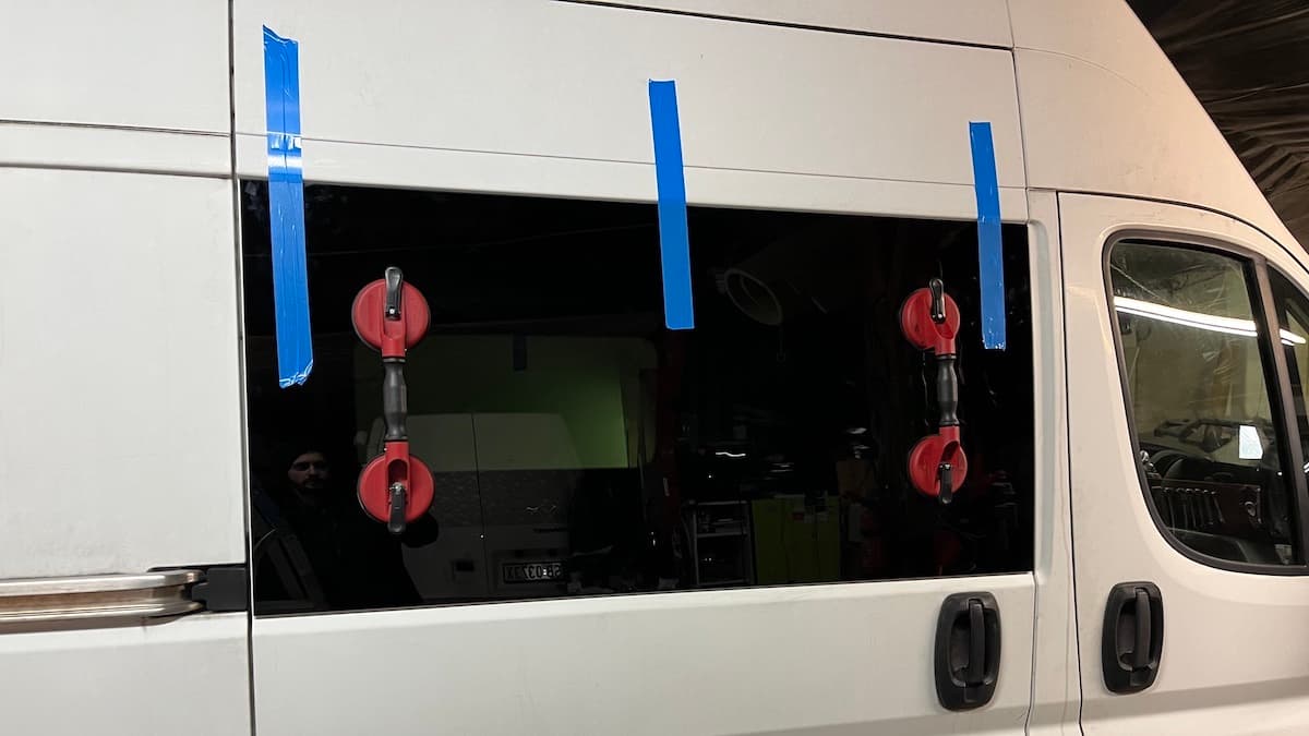 Wymiana szyby bocznej w samochodzie dostawczym. Na szybie zamontowane uchwyty. Szyba zabezpieczona taśmą klejącą.