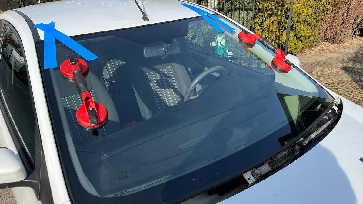 Wymiana szyby czołowej w samochodzie osobowym u klienta. Na szybie są uchwyty do trzymania. Szyba zabezpieczona taśmą klejącą do dachu.